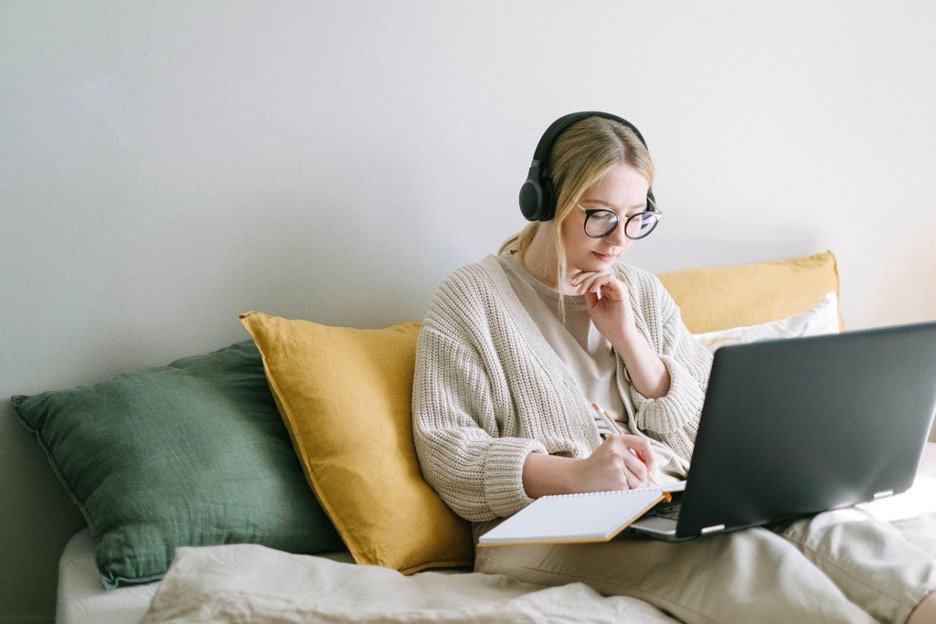 Woman sat on a sofa on a laptop with headphones on writing in a book