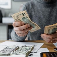 man in a grey sweater jumper counting money with a calculator & pen on desk