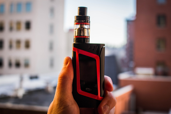 Hand holding a black vape machine with a red outline on the side with faded out background of a building