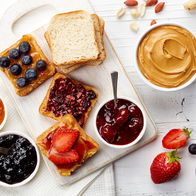 A selection of healthy snack options, including bread with peanut butter, various fruit jams, blueberries, strawberries, and nuts, arranged on a white wooden board